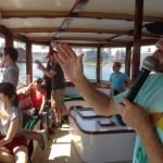 A man in the foreground is holding a microphone and gesturing with his arm as several people in the foreground look out from the inside of a boat to the waterfront