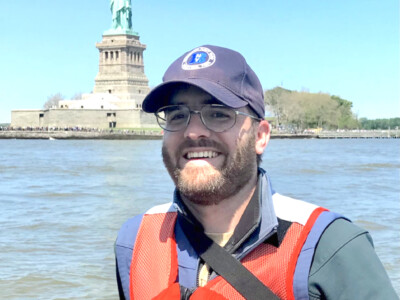 Andrew Gustafson wearing a blue baseball cap and an orange life jacket on a boat in front of the Statue of Liberty