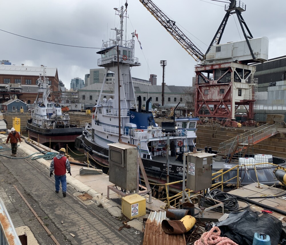 Workers walk alongside a large stone dry dock in the Brooklyn Navy Yard with two white tugboats below and a red and white crane on the right.