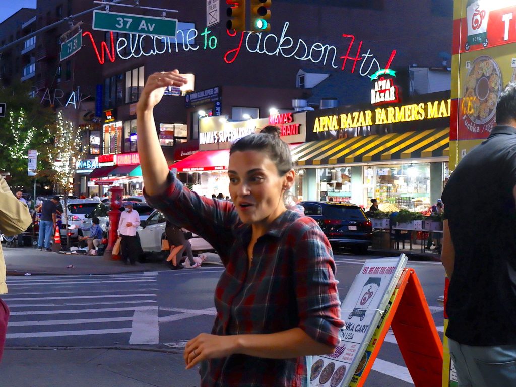 Amanda, a woman wearing a plaid shirt speaking to a group with one hand over her head, and a lighted sign over the street behind her that says "Welcome to Jackson Heights"