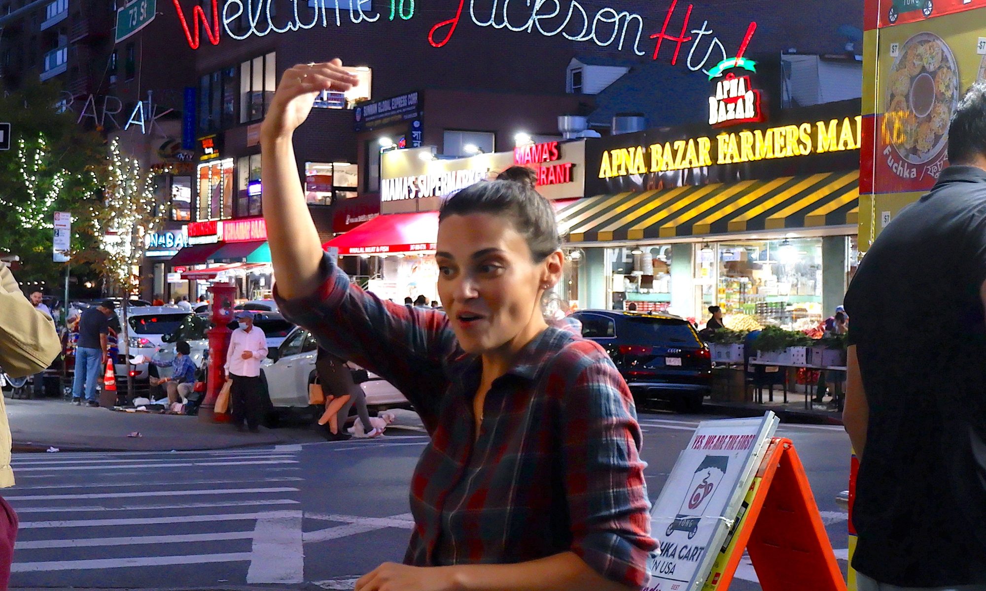 Amanda, a woman wearing a plaid shirt speaking to a group with one hand over her head, and a lighted sign over the street behind her that says "Welcome to Jackson Heights"