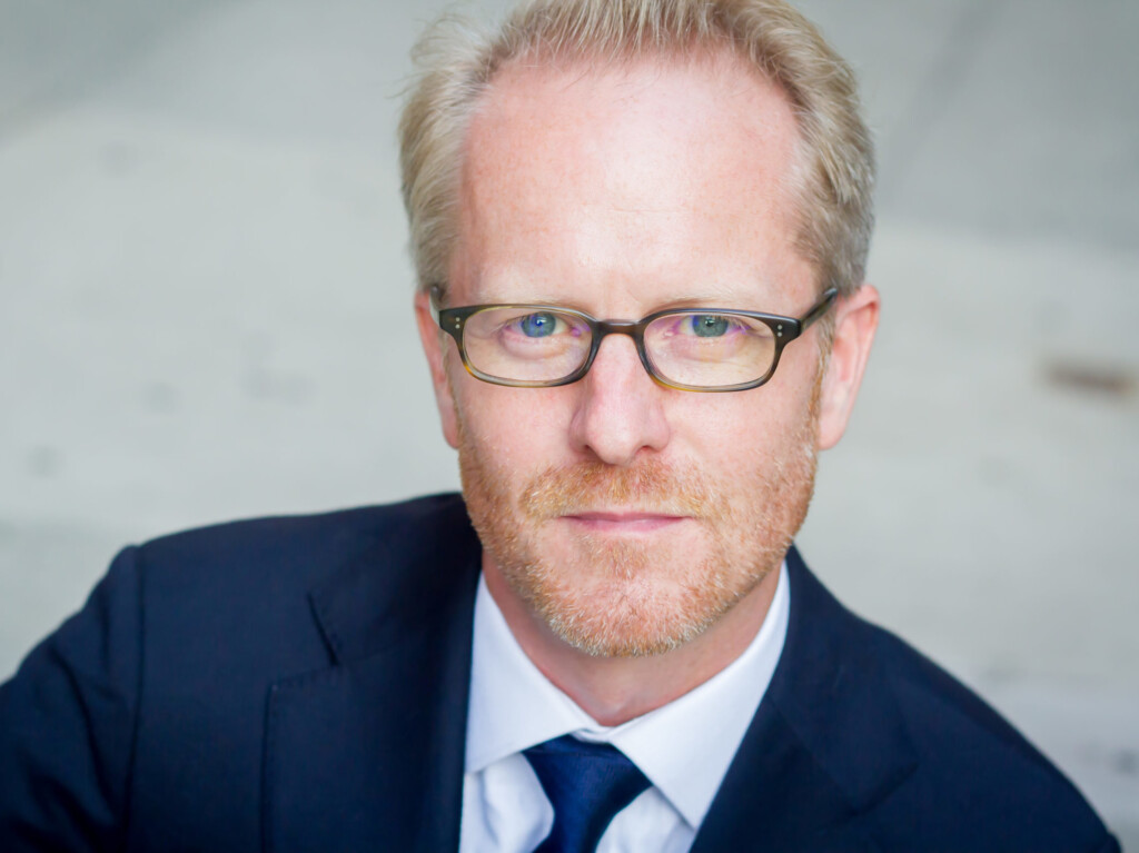 Headshot of Doug, a man with blonde hair, glasses, and a light beard, wearing a suit.