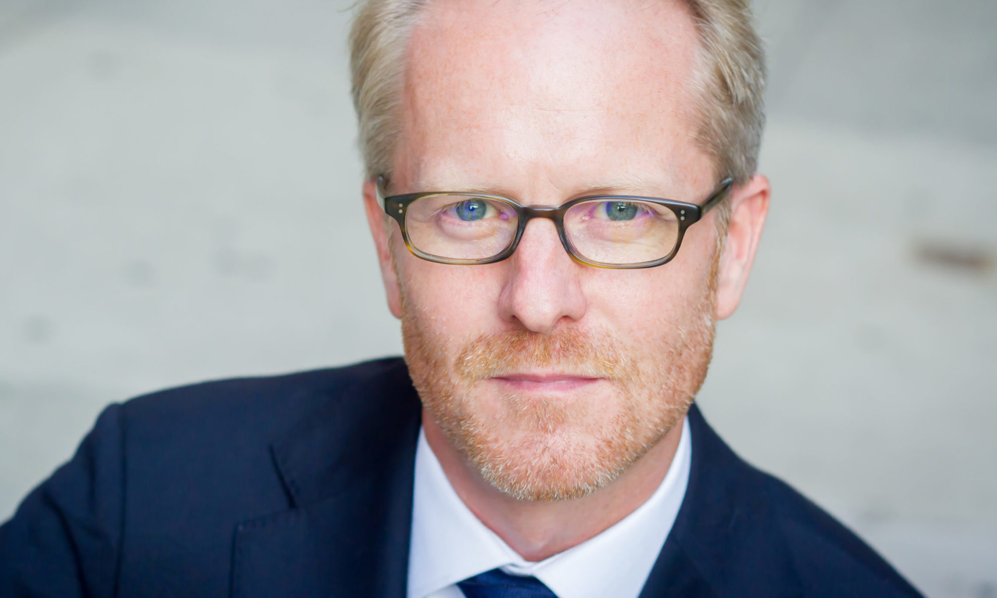 Headshot of Doug, a man with blonde hair, glasses, and a light beard, wearing a suit.