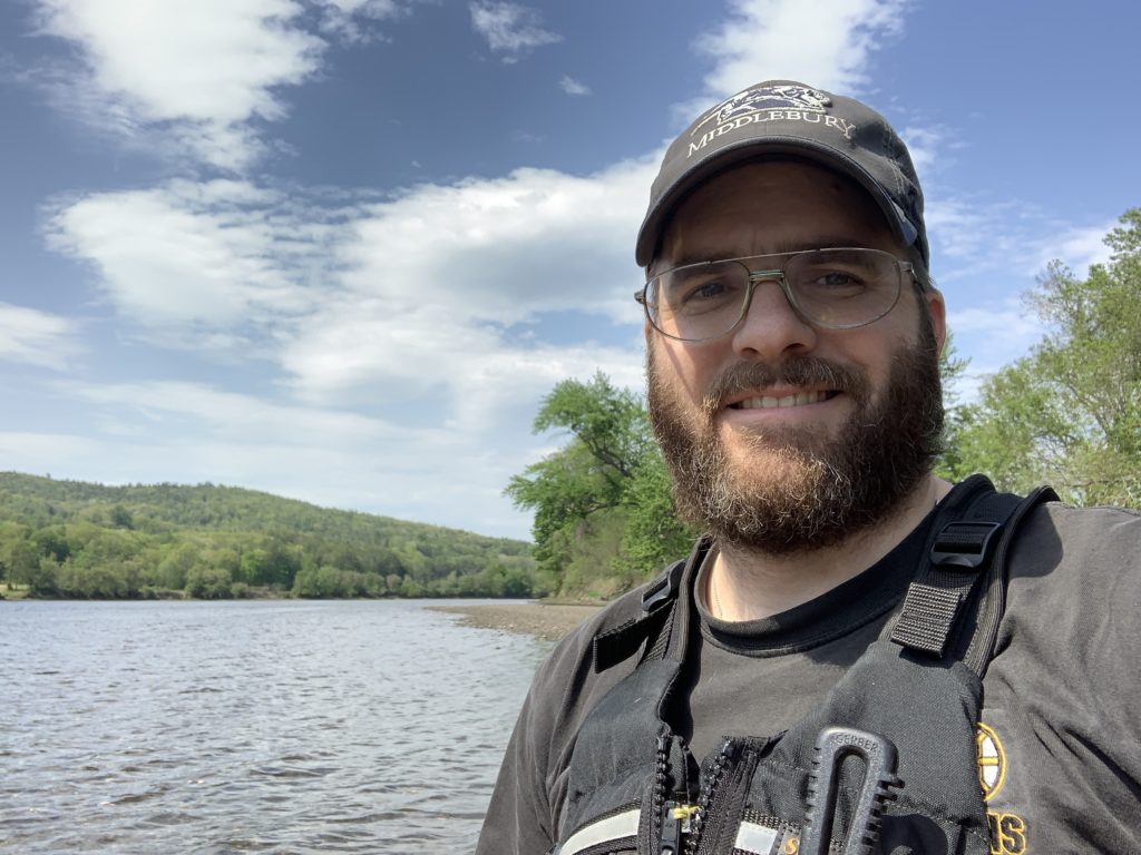 Andrew, a man with a beard wearing a baseball cap and a life jacket, with a river and forest behind him.