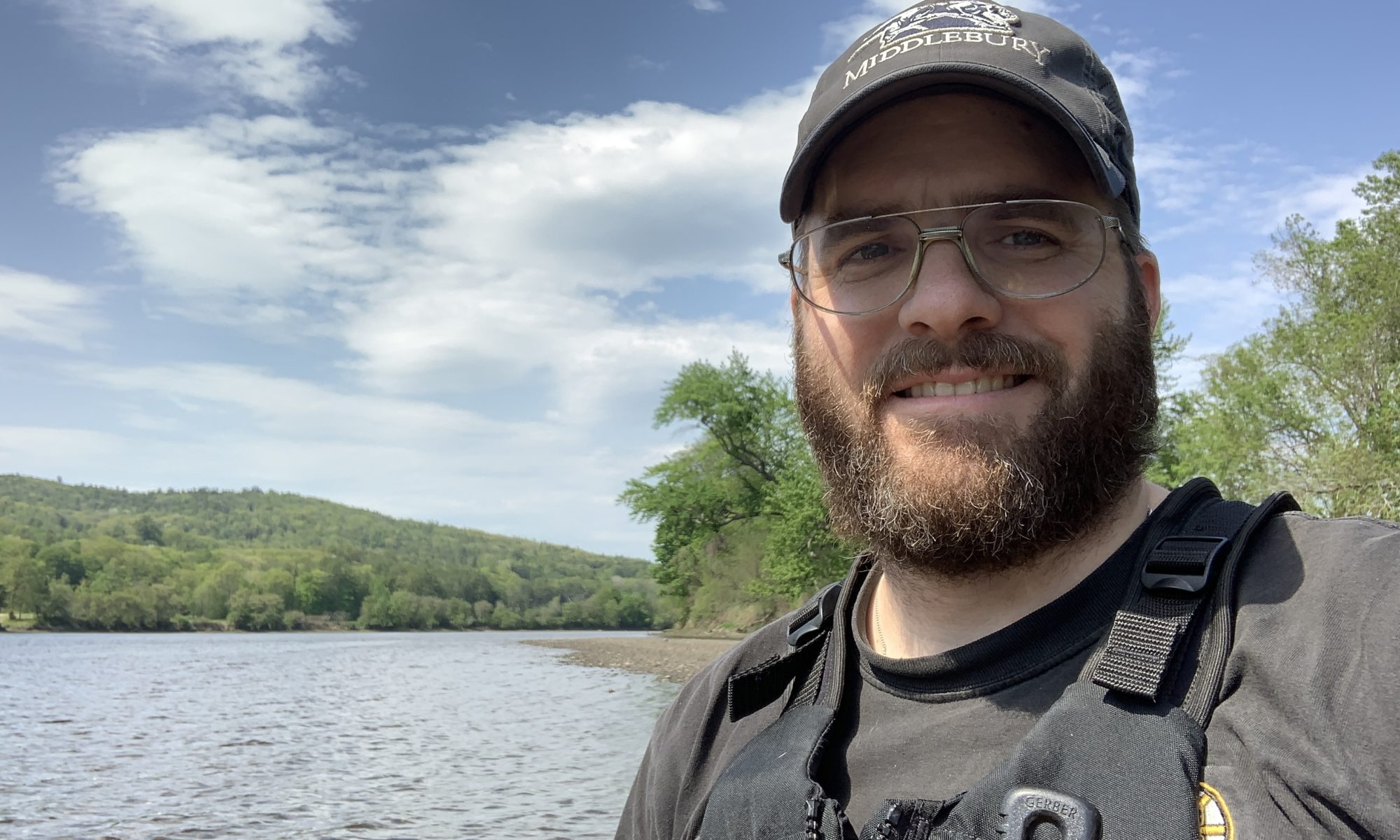 Andrew, a man with a beard wearing a baseball cap and a life jacket, with a river and forest behind him.