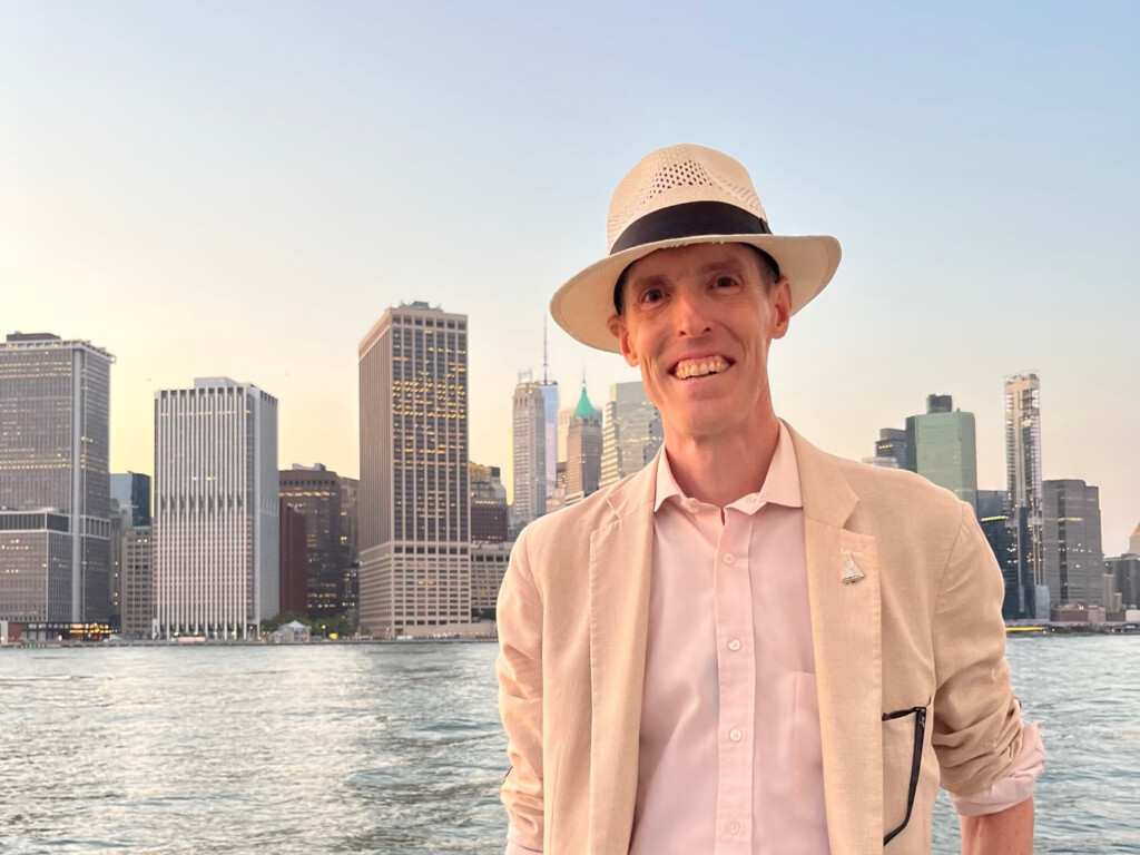 Stefan, a man wearing a pink shirt, light jacket, and Panama hat, standing on a boat in the East River in front of the Manhattan skyline.