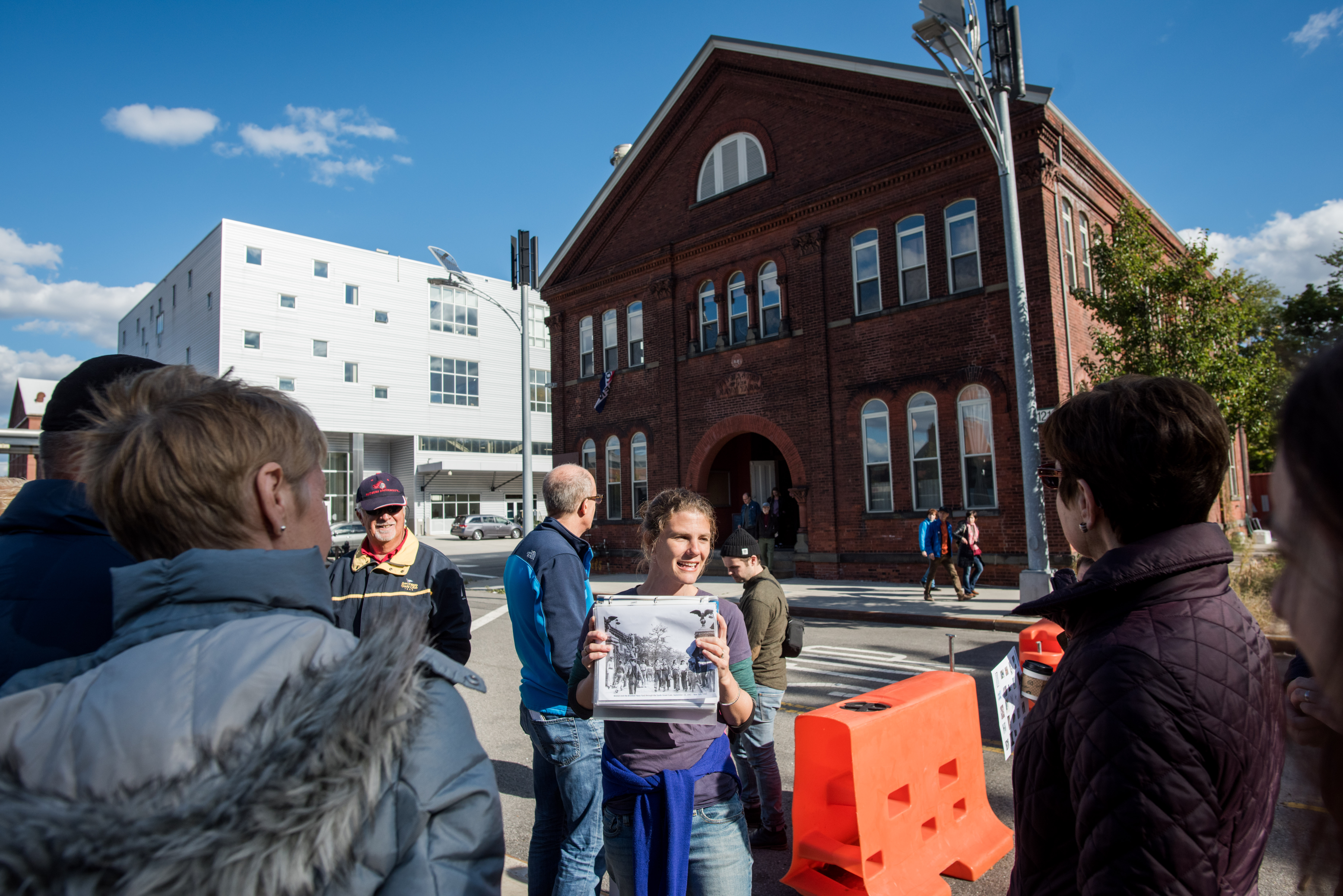 A guide is smiling and holding a historical photo of workers walking into the entrance of the Brooklyn Navy Yard as people look on. In the background, there are wind and solar-powered street lamps, a historic red brick building, and a modern industrial multi-story building.