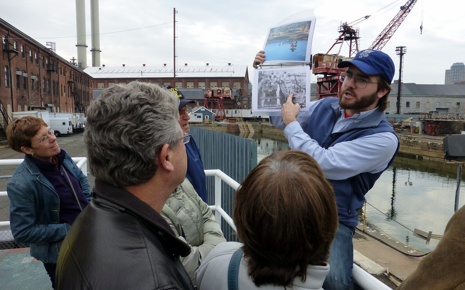 A tour guide points at a black and white photo of two ships as guests look on. They are on a platform that looks out onto a water-filled dry dock that has a red rusty crane next to it.