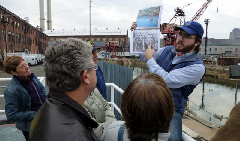 A tour guide points at a black and white photo of two ships as guests look on. They are on a platform that looks out onto a water-filled dry dock that has a red rusty crane next to it.