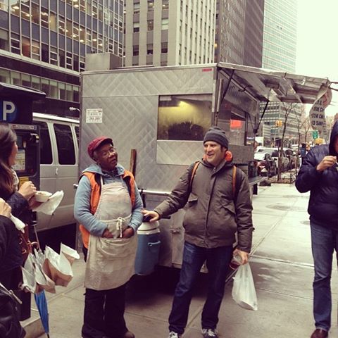 Veronica Julien stands in front of her metal food cart on Water Street and talks to a tour guide and tour group as they smile. wearing glasses in an apron and a young man next to her are both standing in front of a metal food cart on a busy street.