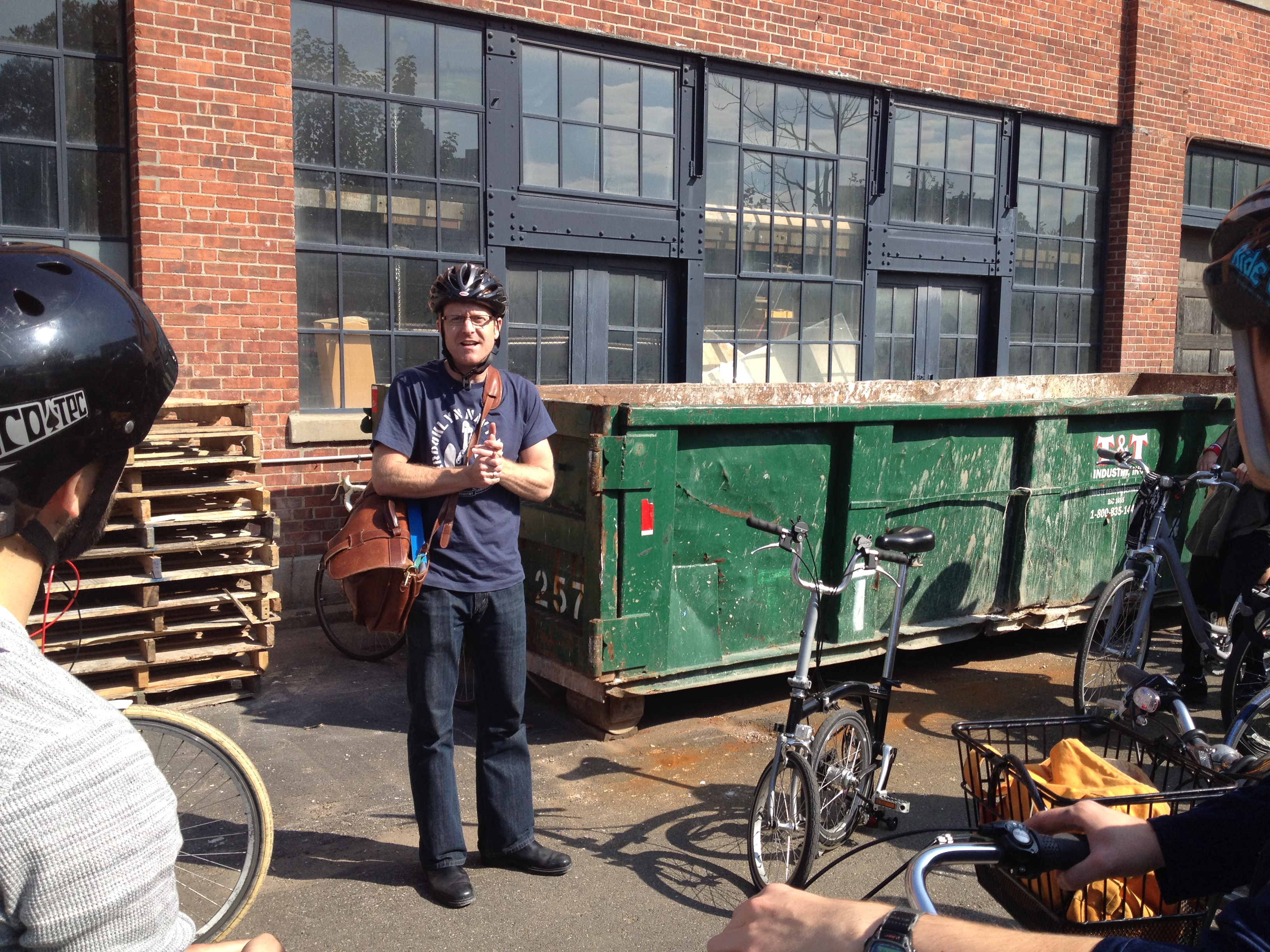 Doug Chapman standing in front of the IceStone factory and a dumpster addressing a tour group on a bicycle tour.
