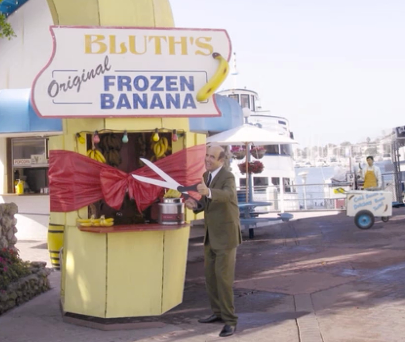 Still from Arrested Development, George Bluth with gian scissors cutting a red ribbon on a Bluth's Original Frozen Banana stand.