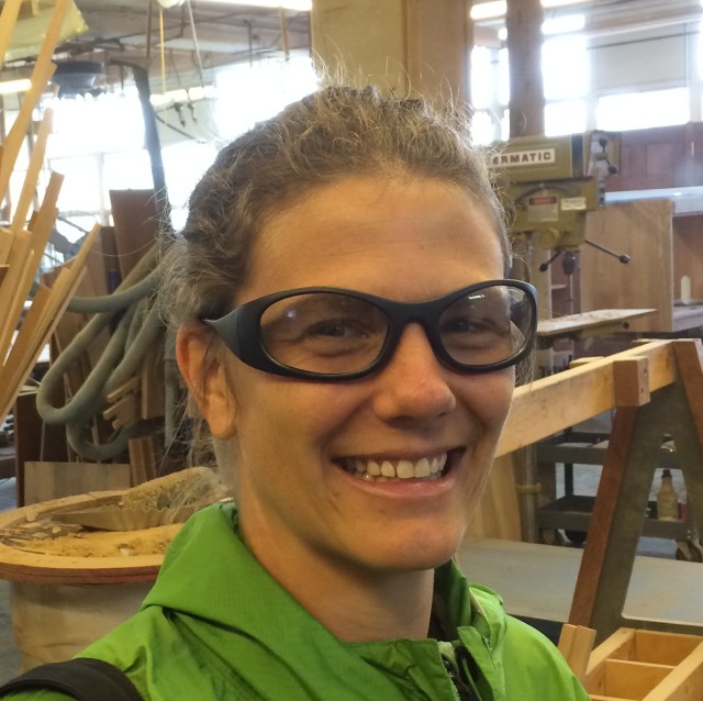Cindy wearing safety goggles and smiling at the camera with a woodworking shop in the background