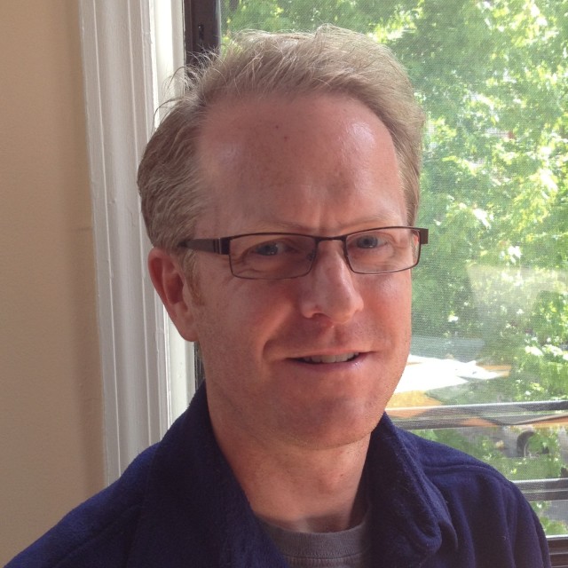 Doug wearing a blue collared shirt and rectangular thin-framed glasses looks at the camera and smiles in front of a window with a tree in the background