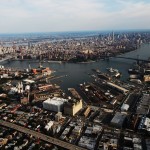 Aerial perspective of the Brooklyn Navy Yard with industrial buildings and dry docks along Wallabout Bay. Manhattan is in the background.