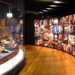 A museum exhibit that includes a series of photographs and videos to the right and an exhibit case with commercial products to the left