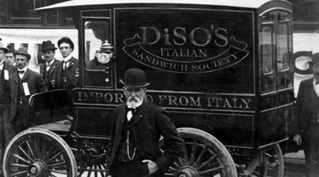 Black and white photo depicting a man in a suit and black hat standing in front of a food truck that reads, "Diso's Italian Sandwich Society" and "Imported from Italy"