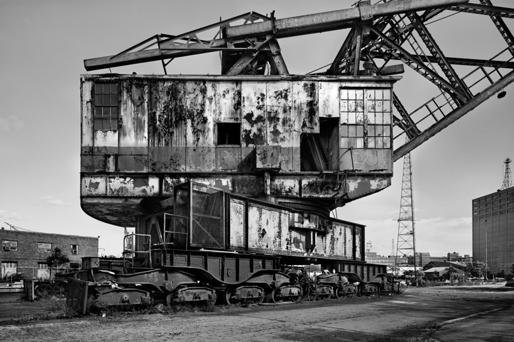 Black and white photo of an old crane no longer in use set against the backdrop of an industrial building in the distance