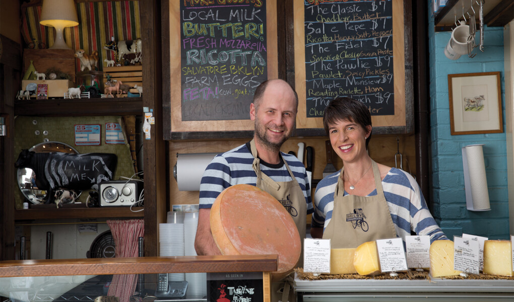 Co-owners - a man and a woman - of Saxelby Cheesemongers, both in striped shirts, stand behind a counter that is filled with cheeses on display. There is a chalkboard in the background that lists out a variety of cheeses.