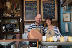 A man and a woman, both in striped shirts, stand behind a counter that is filled with cheeses on display. There is a chalkboard in the background that lists out a variety of cheeses.