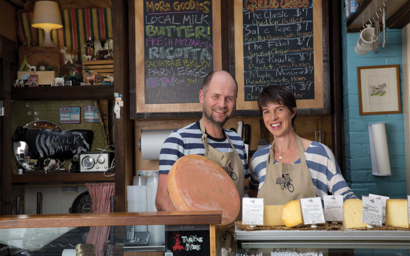 Co-owners - a man and a woman - of Saxelby Cheesemongers, both in striped shirts, stand behind a counter that is filled with cheeses on display. There is a chalkboard in the background that lists out a variety of cheeses.