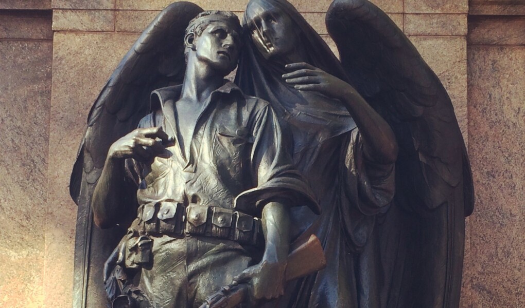 A statue of a soldier who stands clutching his gun and looking off into the distance as an angel begins to wrap her wing and arm around him