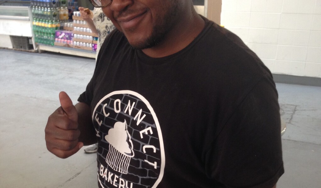 Photo of Daytoine Shaw, a Black man wearing cap, glasses, and t-shirt with Reconnect Bakery written on it, holding a tray of muffins and giving a thumbs up