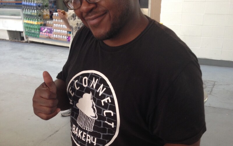 Photo of Daytoine Shaw, a Black man wearing cap, glasses, and t-shirt with Reconnect Bakery written on it, holding a tray of muffins and giving a thumbs up