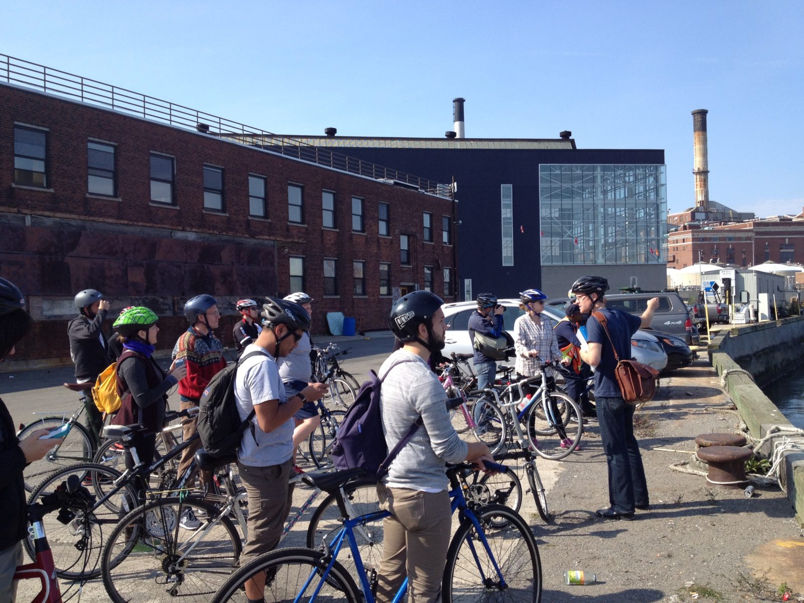 People wearing safety helmets stand with their bicycles at the waterfront by industrial buildings