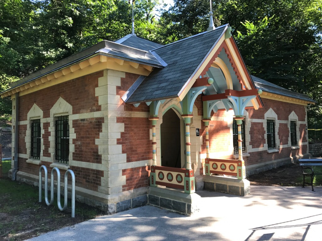 Prospect Park Well House A one-story brick structure with windows and an ornamental portico painted in pastel colors and brown with trees in the background