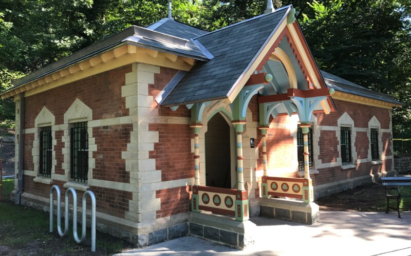 Prospect Park Well House A one-story brick structure with windows and an ornamental portico painted in pastel colors and brown with trees in the background