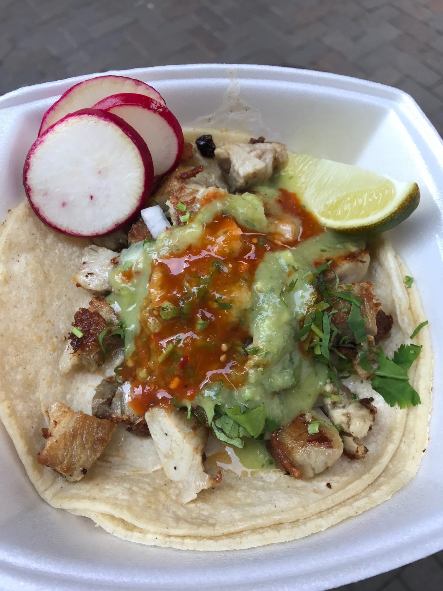 A chicken taco with red and green sauce, cilantro, radishes, and a lime in a styrofoam container