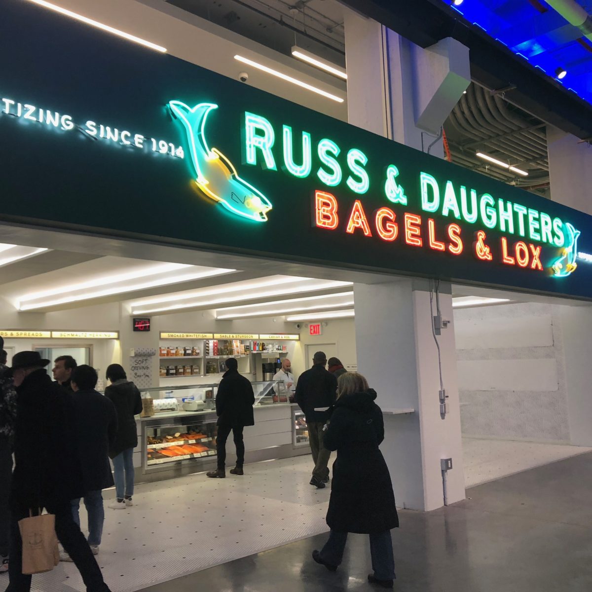 A bright but old-fashioned looking neon sign at a retail shop that has a fish graphics and reads Russ and Daughters Bagels and Lox appetizing since 1914