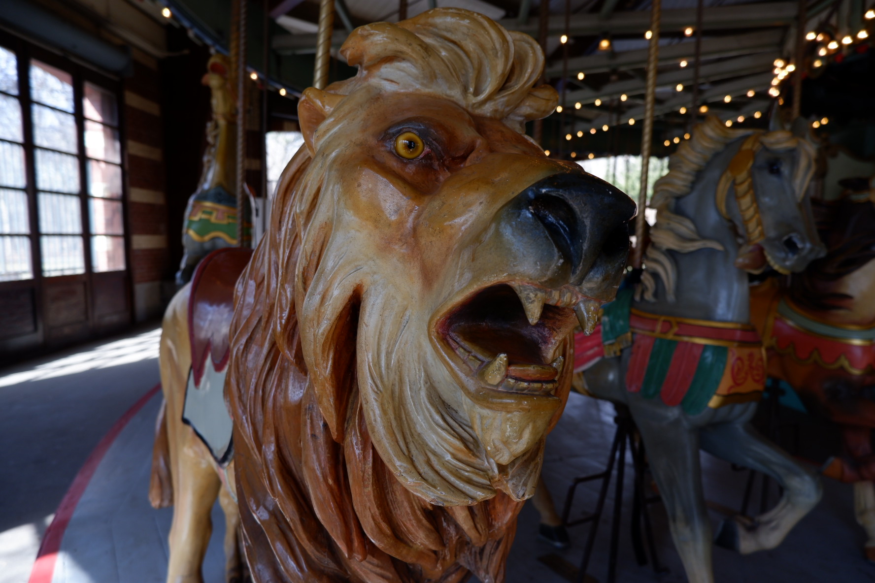 A wooden-carved and painted lion with an open mouth and sharp, fierce teeth and beady yellow and dramatic, aggressive-looking eyes at Prospect Park's carousel