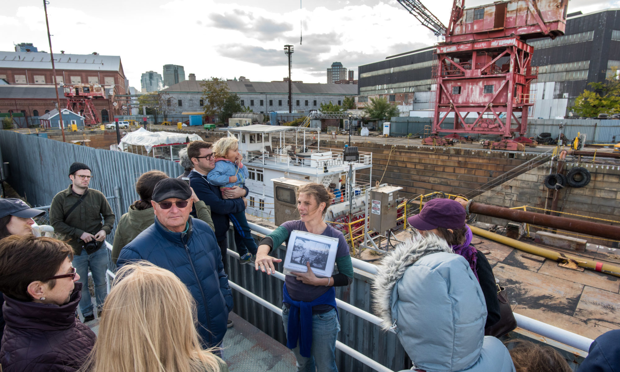 A group of people led by a tour guide stand on a platform looking out at a ferry in a dry dock next to a red crane