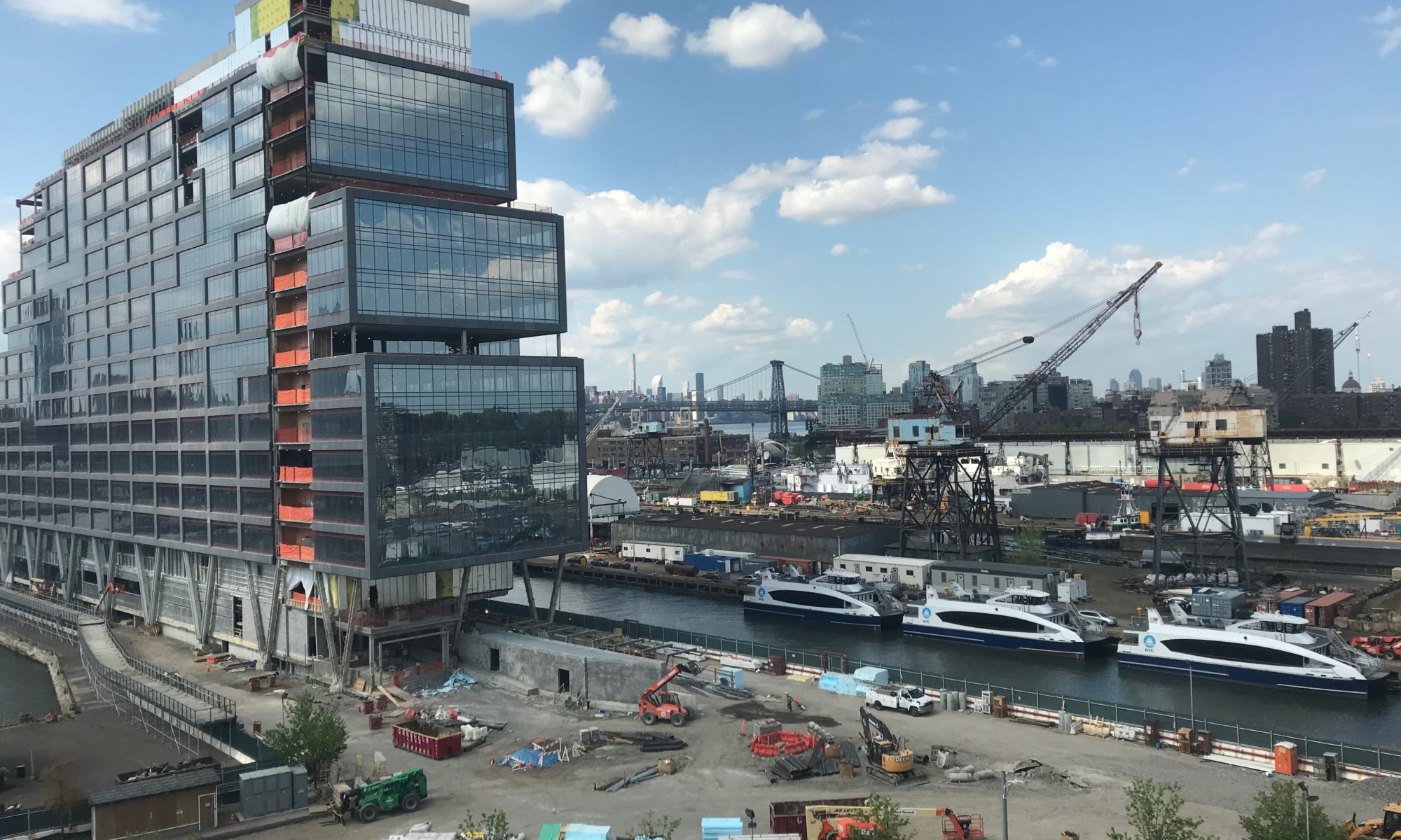 A tall glass office building on a pier next to NYC ferries in a wet berth with ship repair cranes and the Williamsburg Bridge in the background