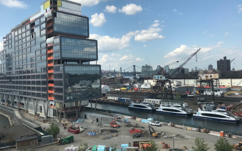 A tall glass office building on a pier next to NYC ferries in a wet berth with ship repair cranes and the Williamsburg Bridge in the background