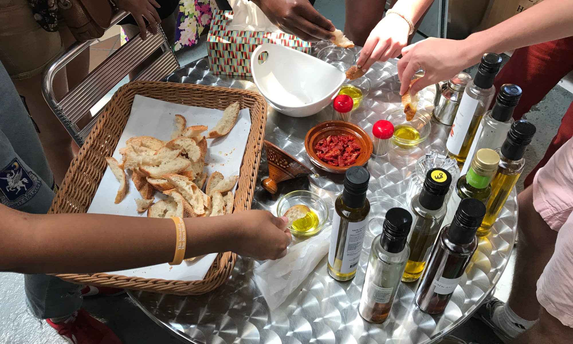 Four hands reaching in from different directions to dip bread into bowls of olive oil on a table with a spread of various olive oils, vinegars, and a bread basket