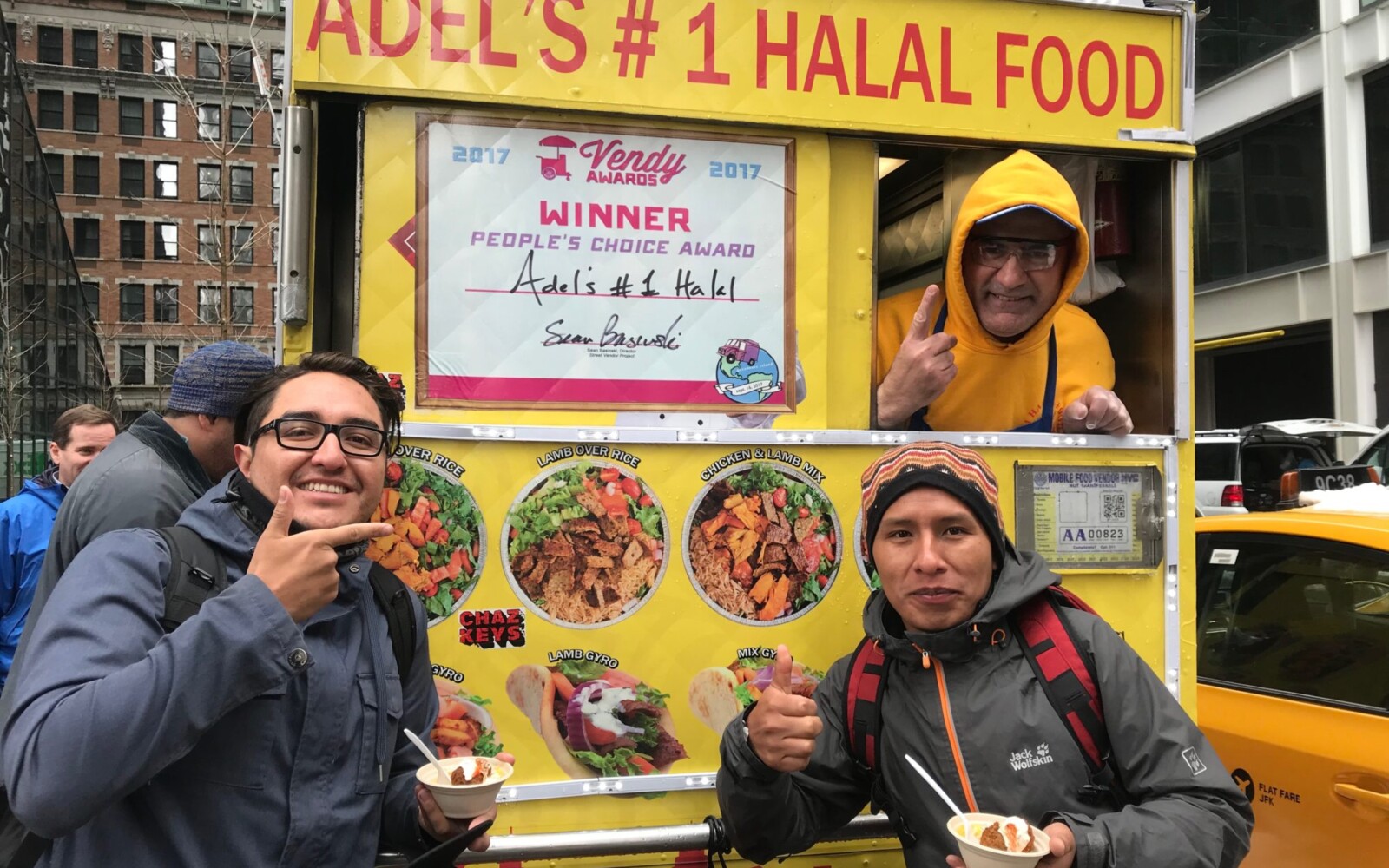 Two young men are holding bowls and posing for the camera in front of a smiling street vendor on a halal food cart