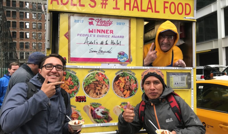 Two young men are holding bowls and posing for the camera in front of a smiling street vendor on a halal food cart