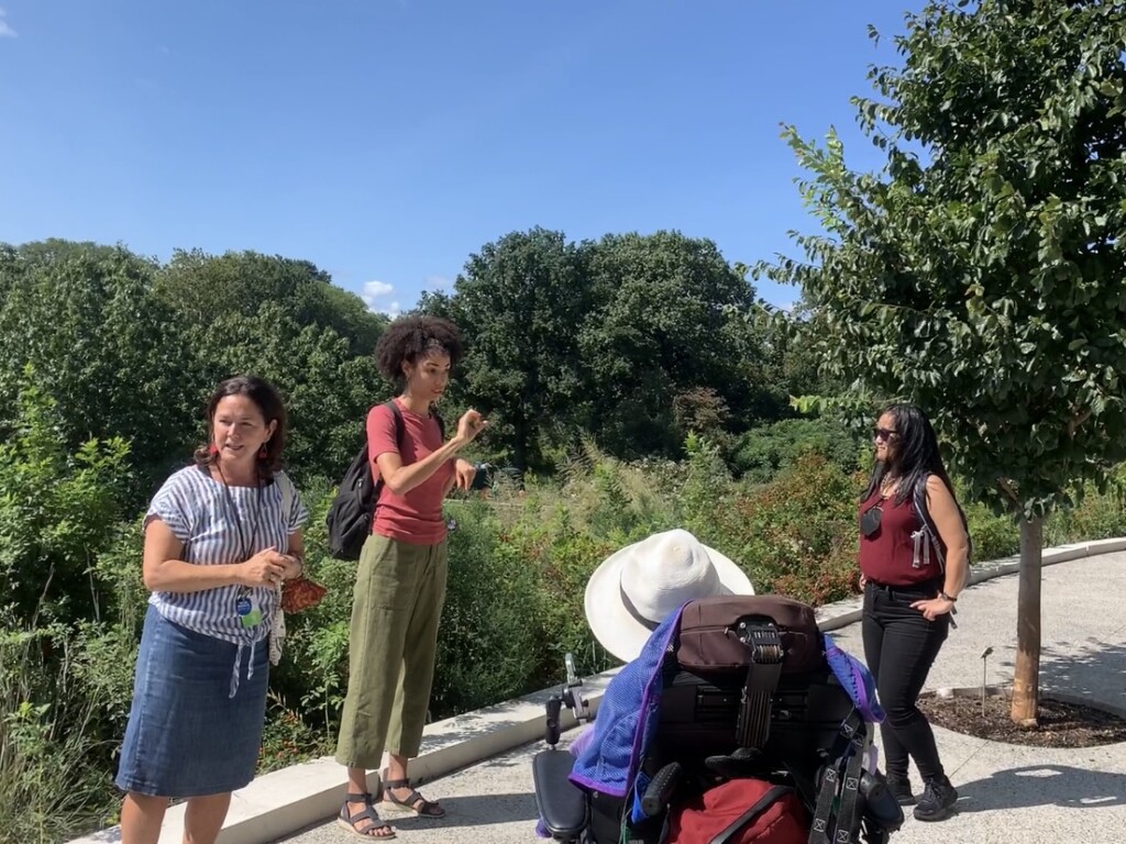 Photo of a tour group showing a woman leading to tour, an ASL interpreter, a guest in a wheelchair, and another guest standing, with trees in the background