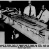 Black and white photo of three workers standing around a wooden model of a ship