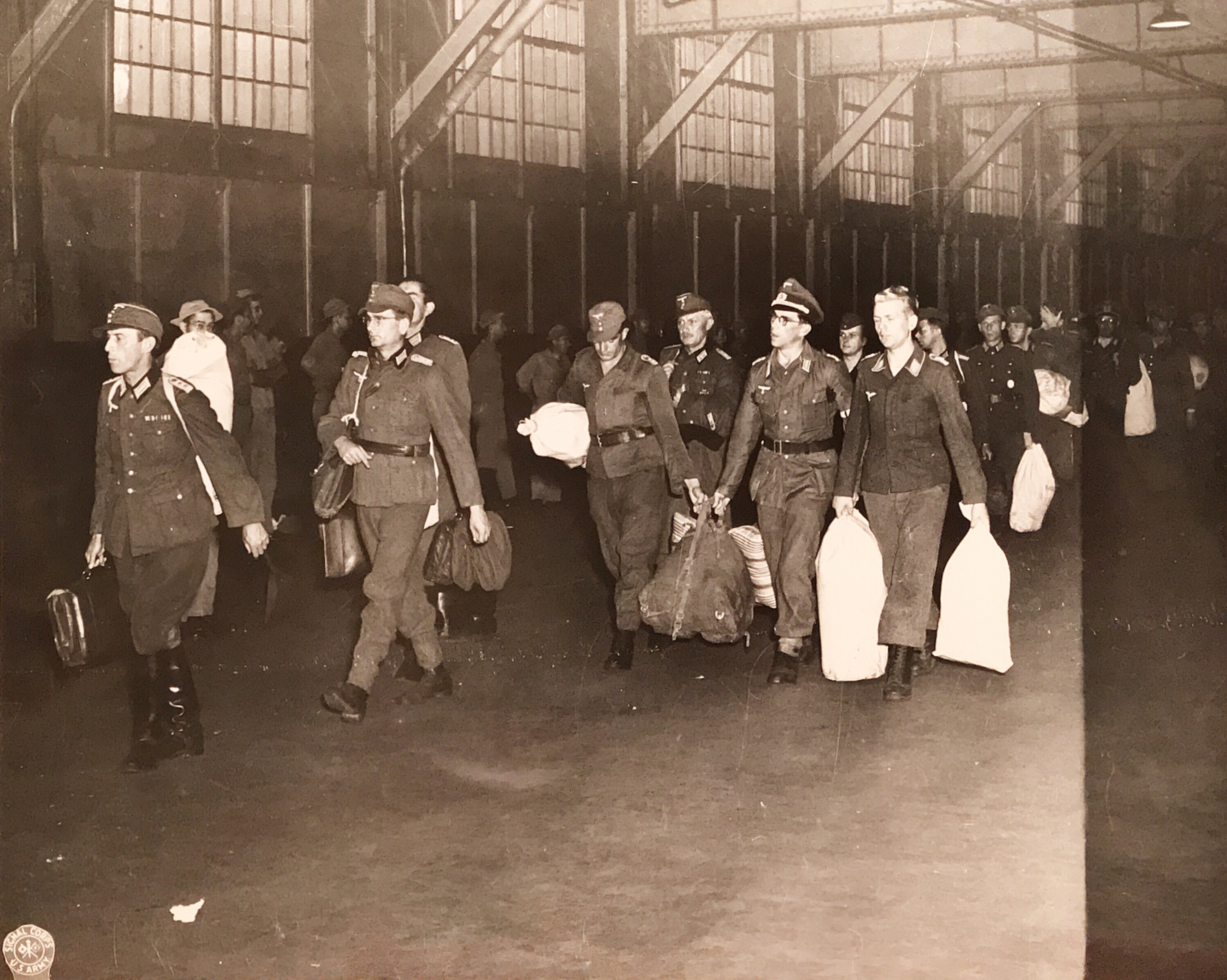 Men in Nazi uniforms carrying duffel bags walk along an indoor pier.