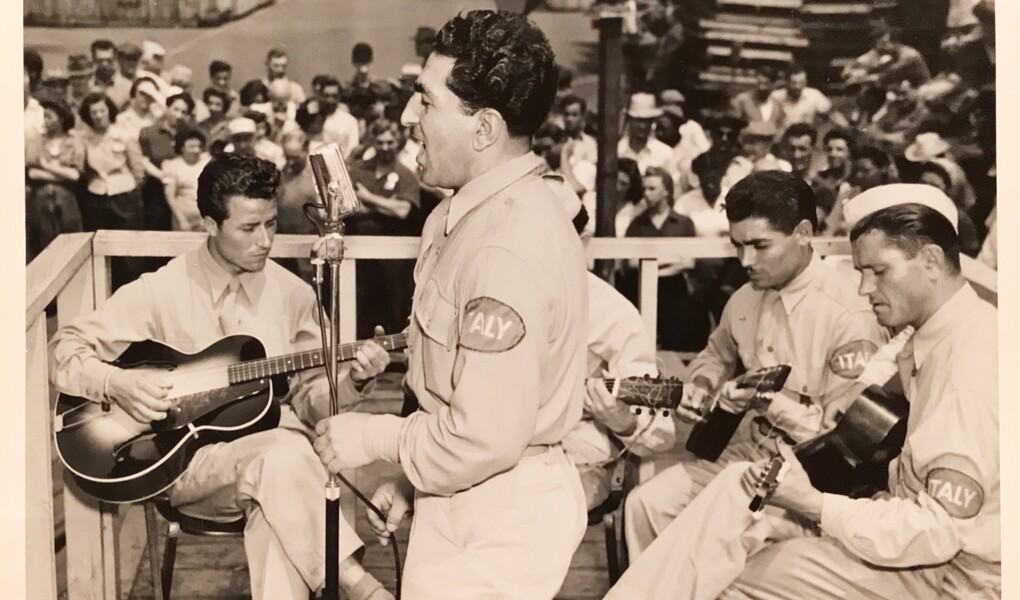 Solider wearing a uniform with "Italy" written on his left arm sings with three guitarists on a bandstand with a crowd in the background.