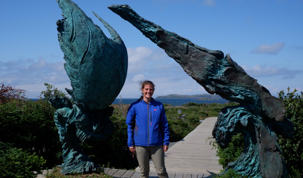 Woman in a blue jacket standing underneath a bronze abstract sculpture with a meadow and ocean in the distance.