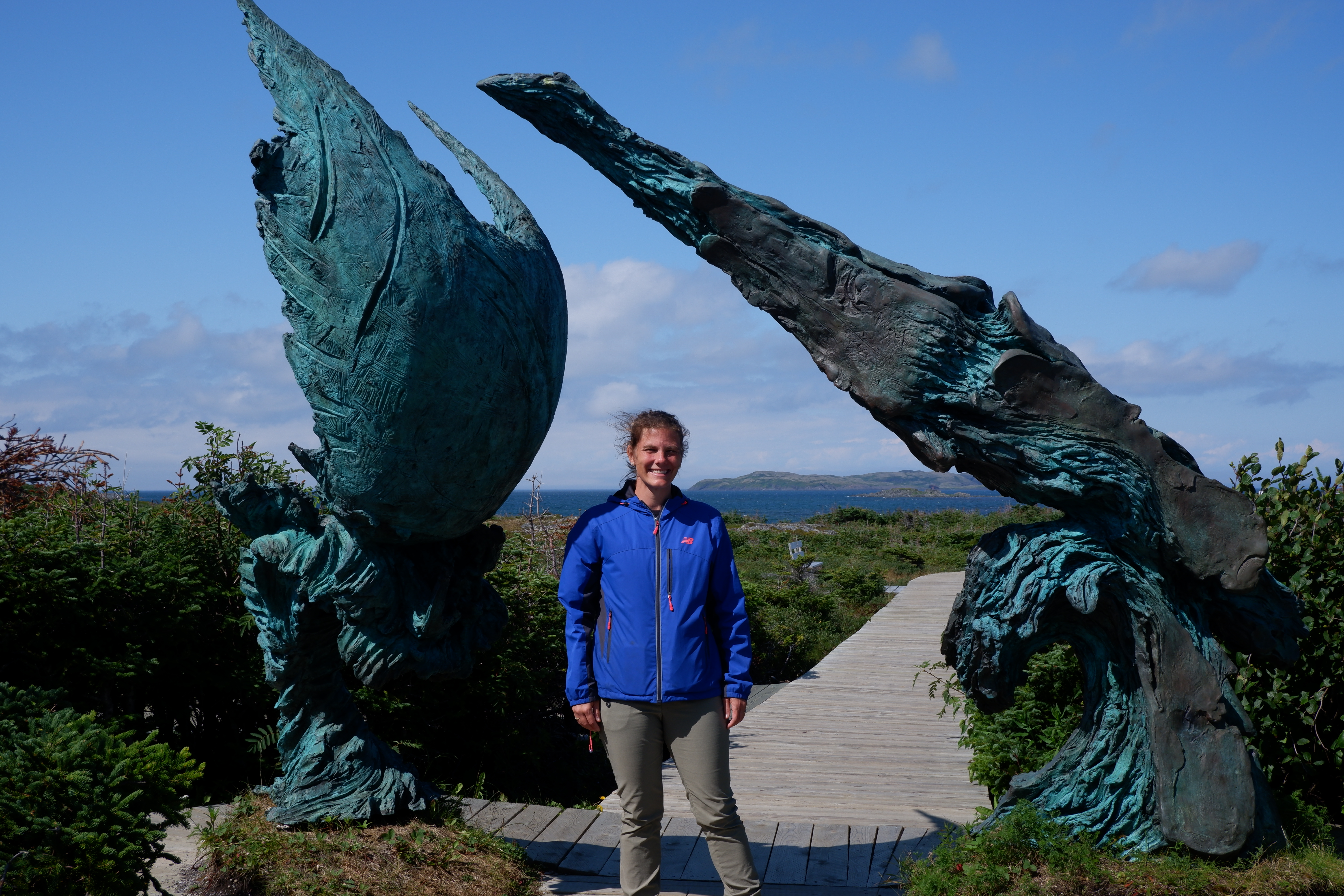 Woman in a blue jacket standing underneath a bronze abstract sculpture with a meadow and ocean in the distance.