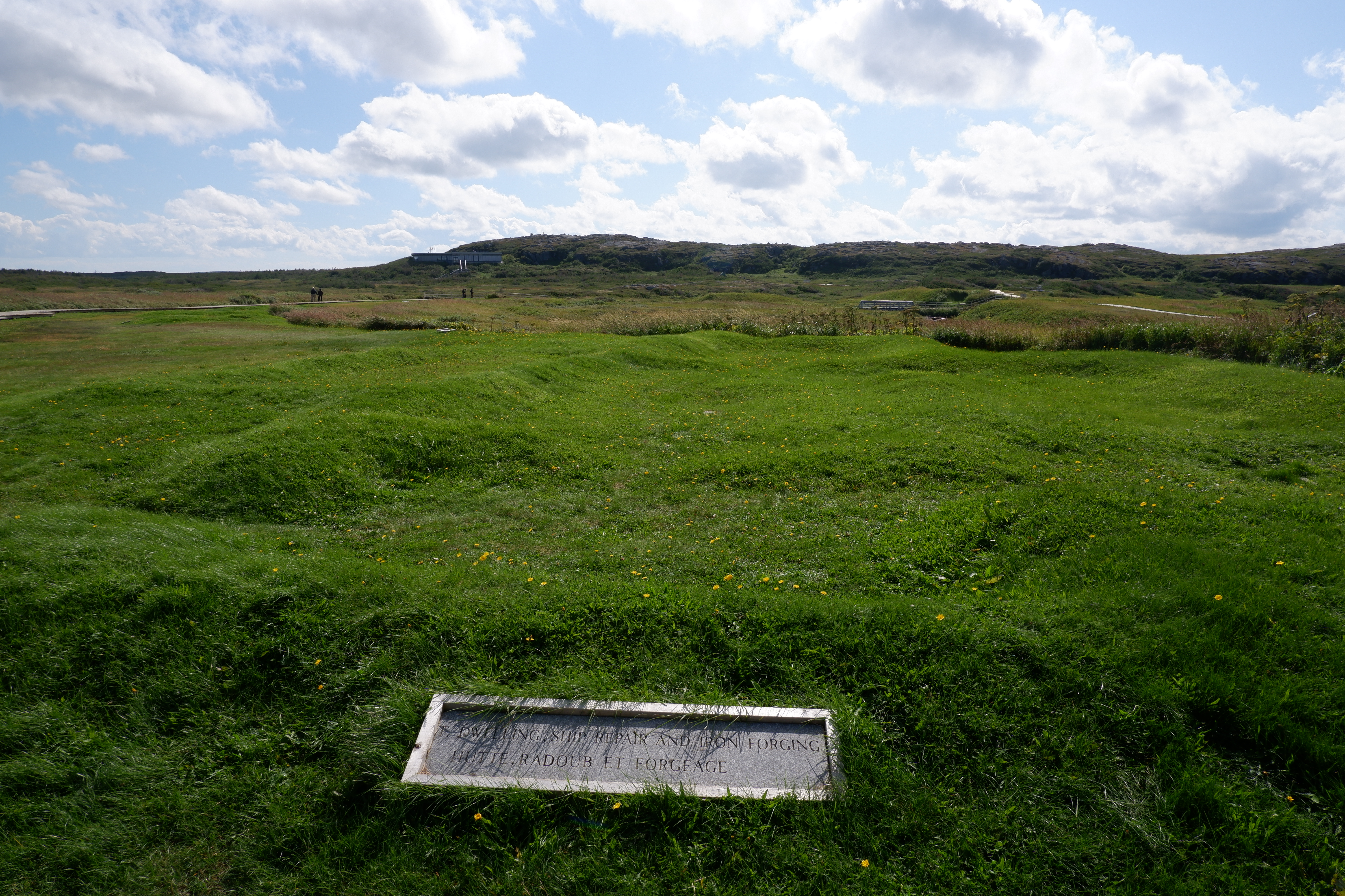 Grassy area with a stone plaque reading "Dwellings, Ship Repair, Iron Forging Hutte Radoub et Forgage."