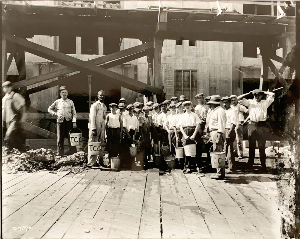 Black and white photo of young boys standing in a construction site holding buckets with scaffolding and the Brooklyn Army Terminal behind them