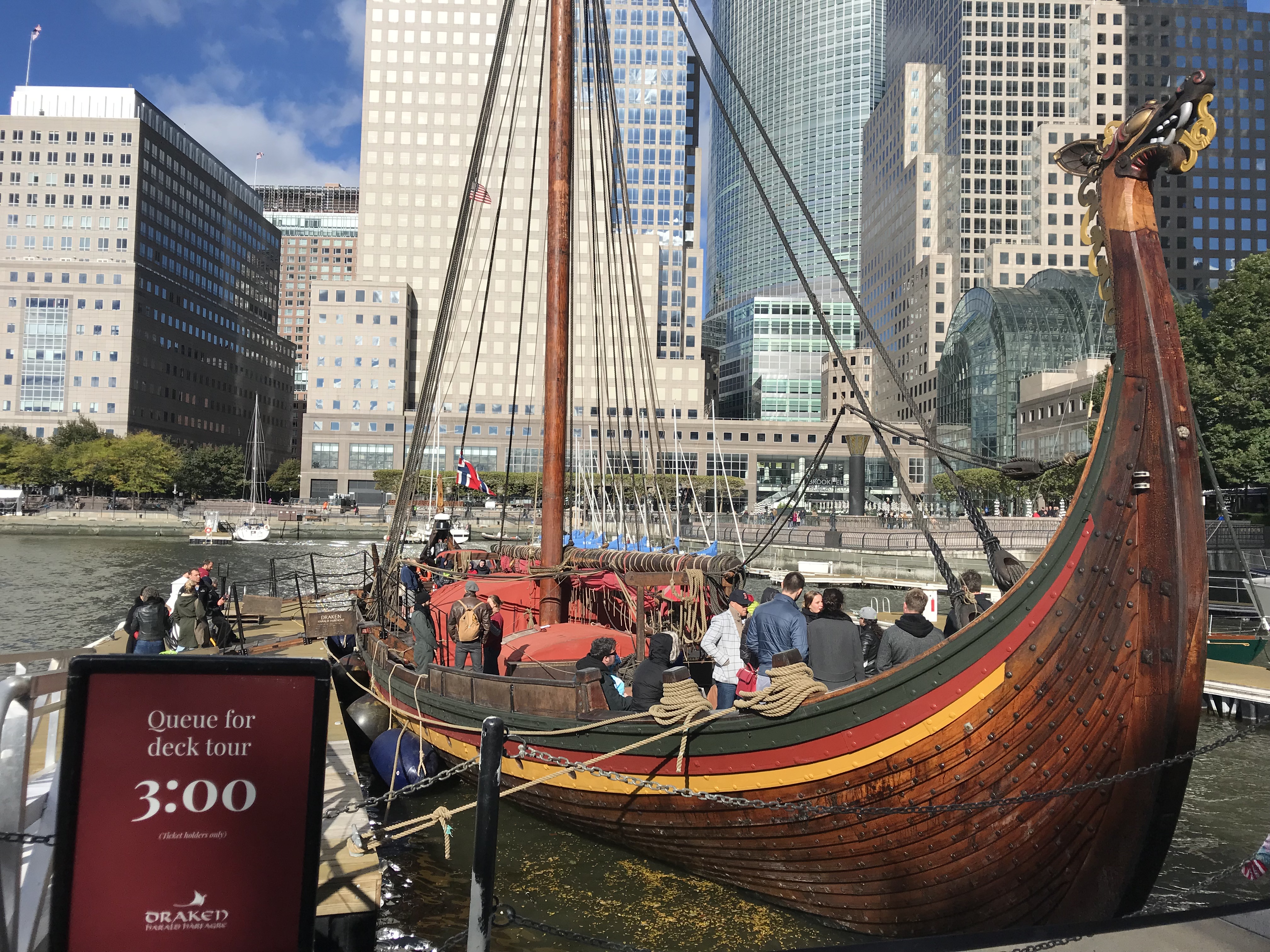 Photo of wooden Viking longship with mast and no sails and tall buildings of Battery Park City in the background.