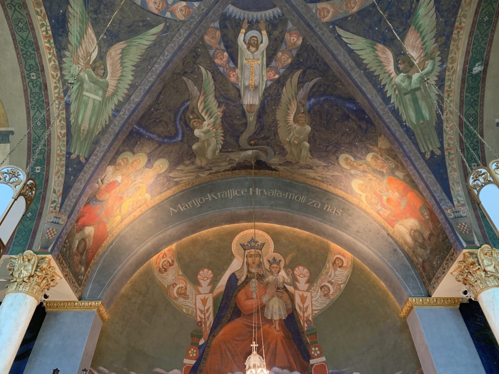 Photo of a church ceiling painted by artist Maxo Vanka depicting Mother Mary wearing a red robe and Baby Jesus on her lap, Jesus ascending to heaven above her